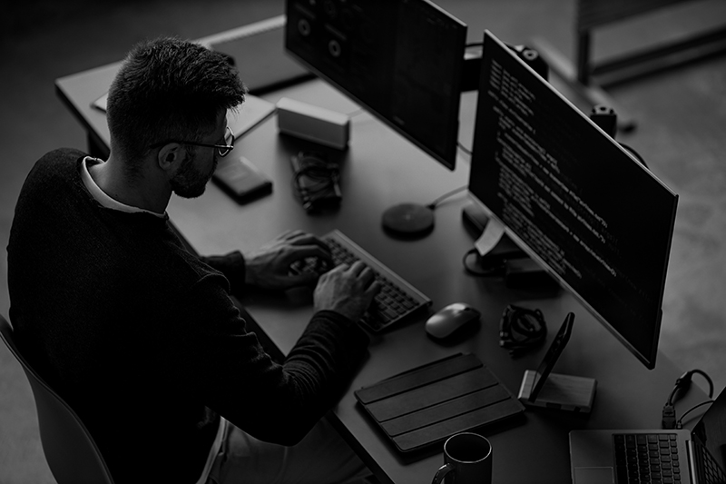 Mitarbeiter arbeitet an mehreren Monitoren mit Programmcode – Symbol Cybersecurity und Softwareentwicklung